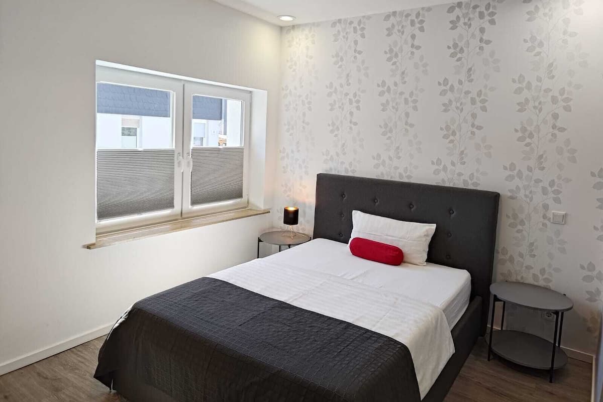 Ein Schlafzimmer mit Bett, Fenster, Nachttisch und einer Wand mit Blumenmuster.