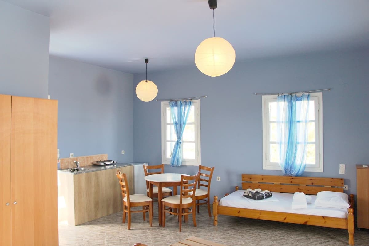 A room with a bed, a dining table with chairs, a small kitchen area, and two windows with blue curtains.