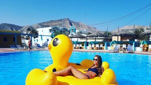 A pool area with a person on a yellow inflatable float, lounge chairs, and a mountain backdrop. - Dodo's Santorini (Santorini)