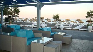A covered outdoor seating area with sofas and tables, overlooking a beach with umbrellas and lounge chairs. - Dodo's Santorini (Santorini)