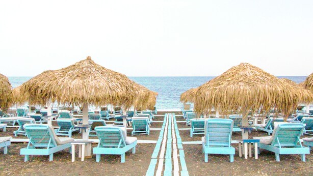 A beach with rows of blue lounge chairs and thatched umbrellas, leading to the ocean. - Dodo's Santorini (Santorini)
