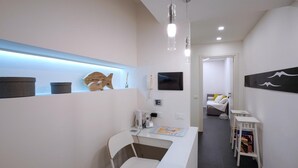 Une chambre d’hôtel moderne avec un bureau blanc, une chaise, une petite étagère ornée d’objets décoratifs, un téléviseur à écran plat et un tableau accroché au mur.