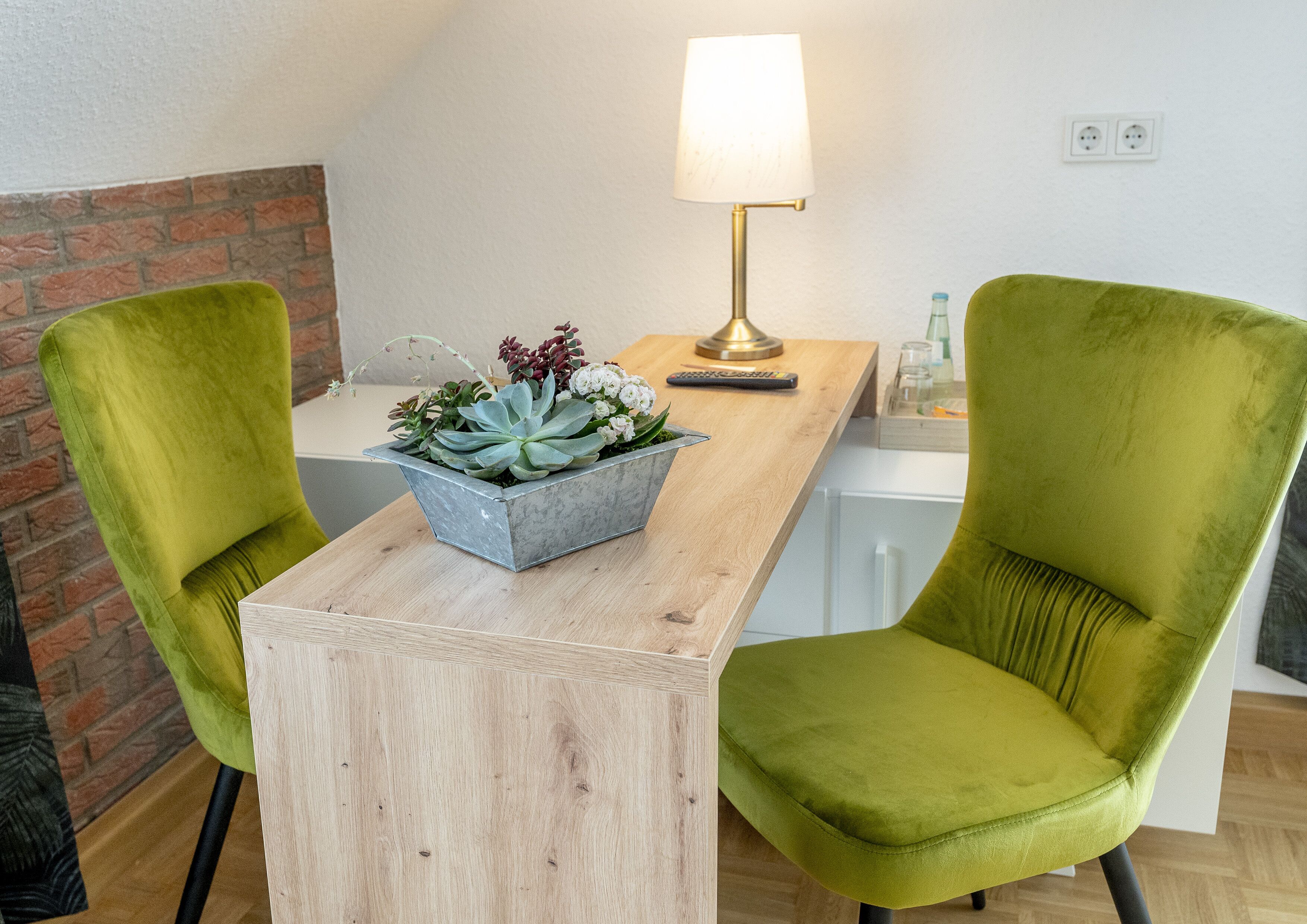 A modern room with a wooden table, two green chairs, a metal planter with succulents, a lamp, and a brick wall.