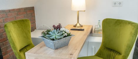 Une pièce moderne avec une table en bois, deux chaises vertes, un pot en métal contenant des plantes succulentes, une lampe et un mur en briques.