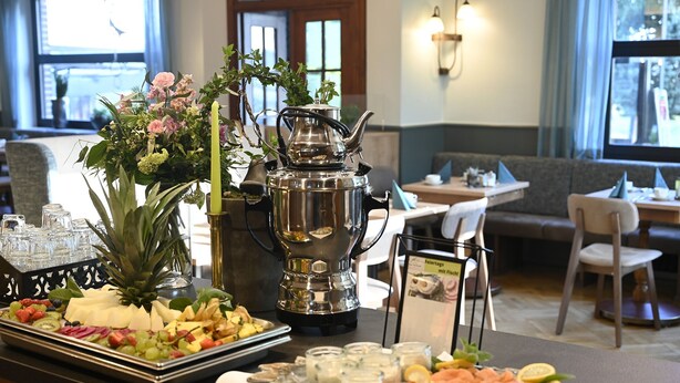 Un espace repas avec une table de buffet proposant une variété de fruits, de fromages et de saumon fumé. On y trouve une théière en argent et un vase rempli de fleurs. La pièce dégage une atmosphère chaleureuse, avec un canapé et une table en arrière-plan.