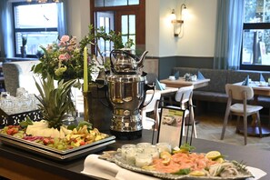 Un espace repas avec une table de buffet proposant une variété de fruits, de fromages et de saumon fumé. On y trouve une théière en argent et un vase rempli de fleurs. La pièce dégage une atmosphère chaleureuse, avec un canapé et une table en arrière-plan.