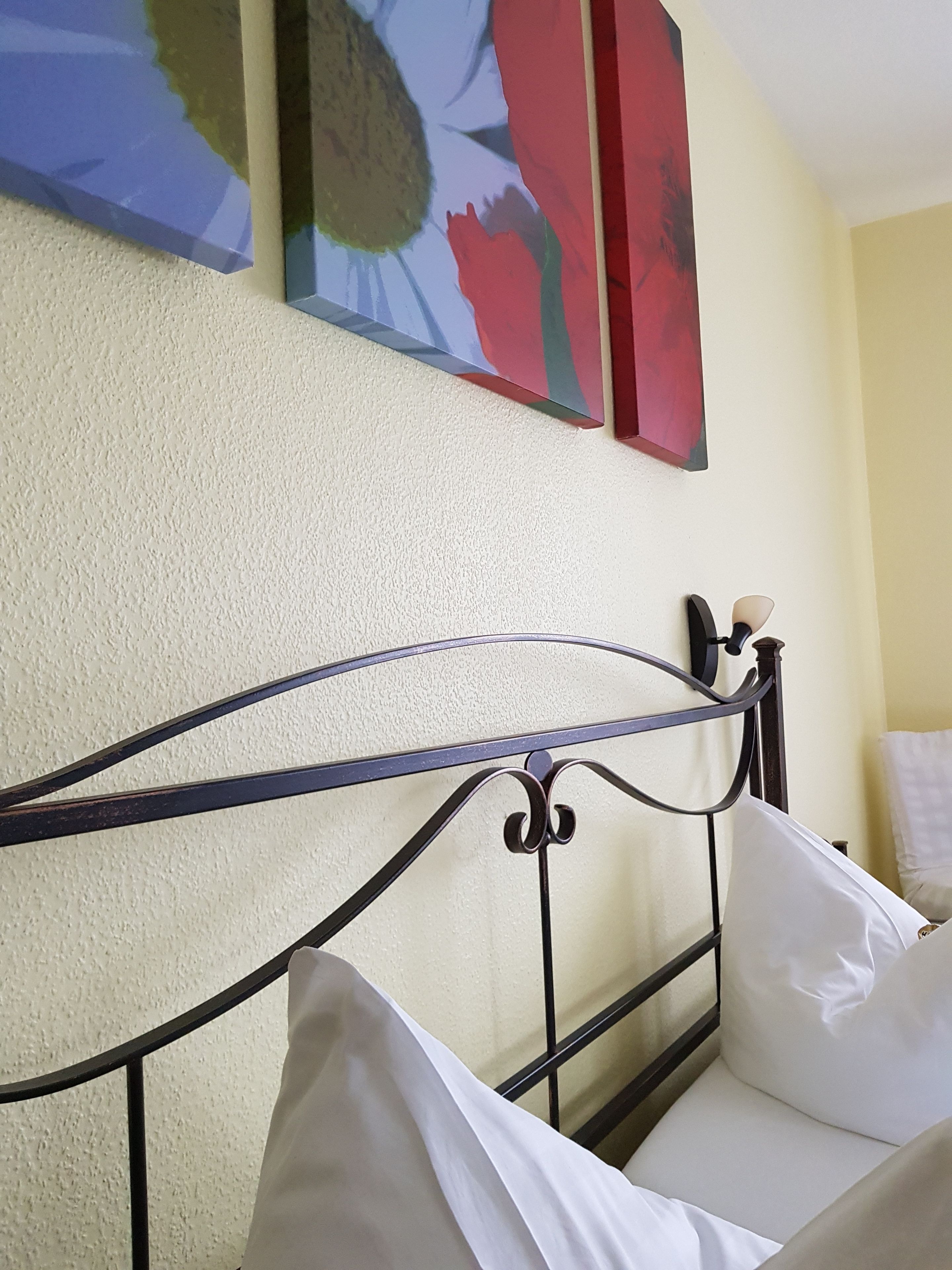 Una cama con ropa de cama blanca y cabecero de metal en una habitación con paredes amarillas y un cuadro de flores.