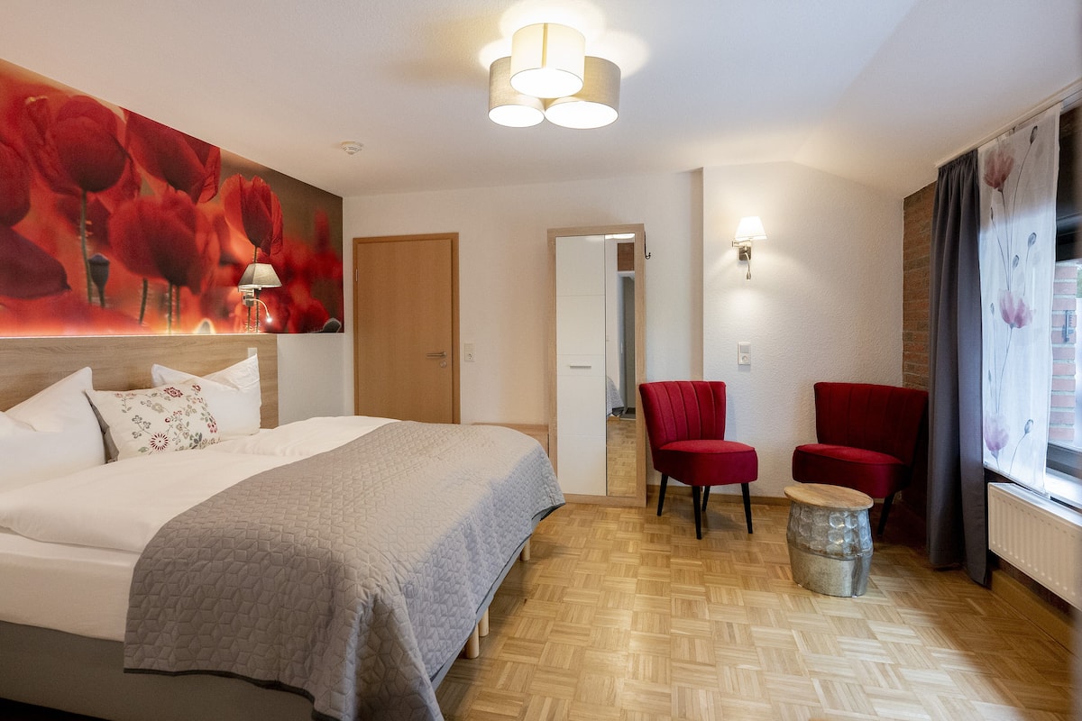 Habitació d'hotel amb un llit, dues cadires vermelles, una taula rodona petita, un mirall i una finestra amb cortines.