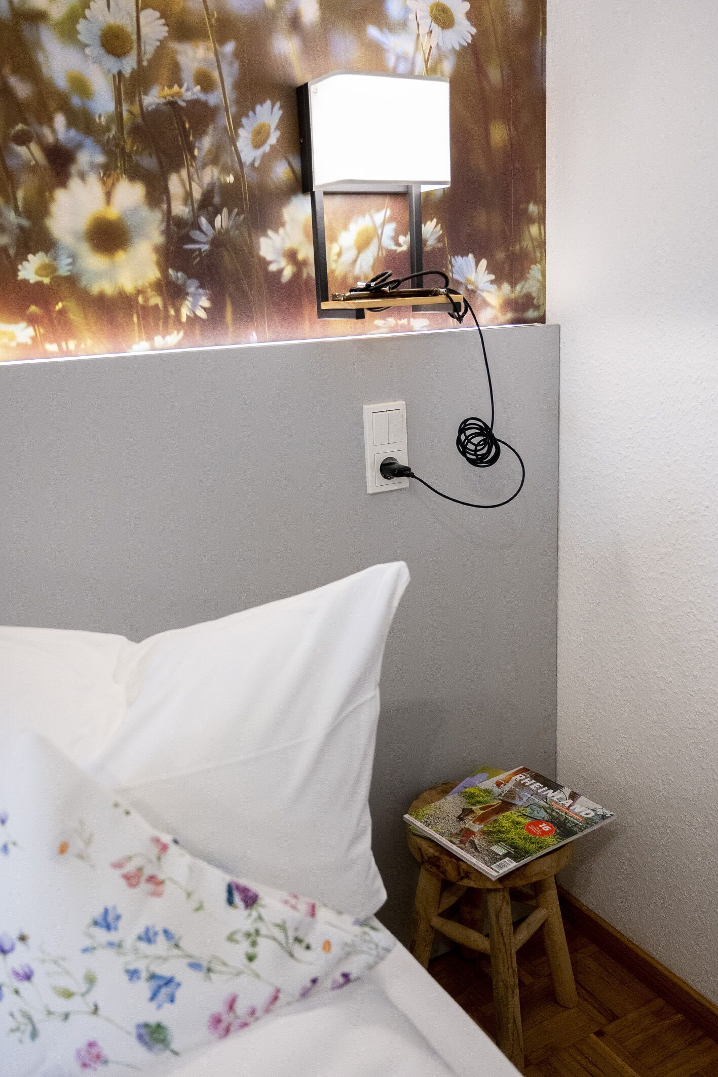 Un dormitorio con una cama, una mesita de noche con una revista, una lámpara de pared y un papel tapiz floral.