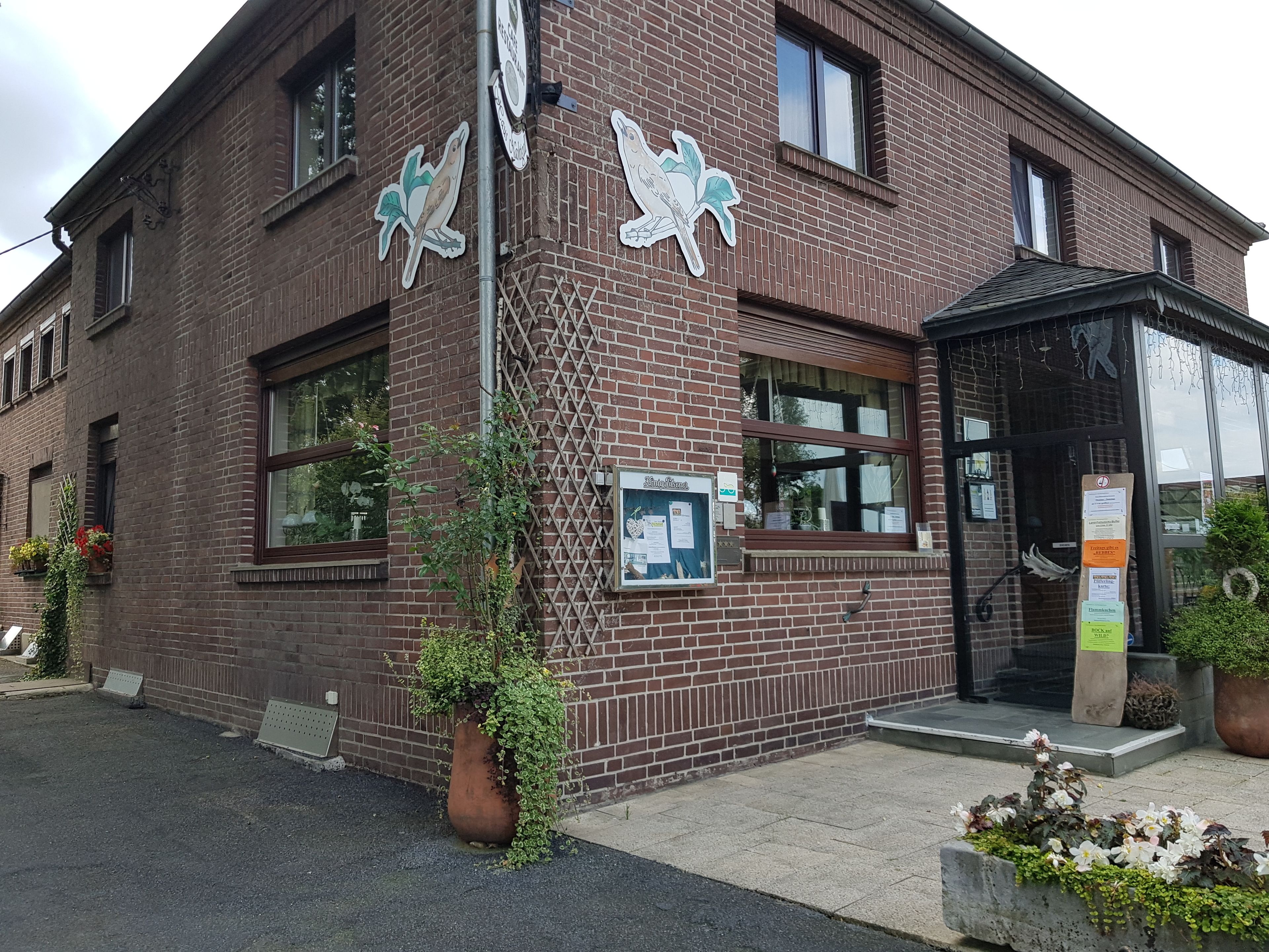 Ein Gebäude aus Ziegelsteinen mit großen Fenstern und einer Tür, umgeben von Topfpflanzen und einer Bank.