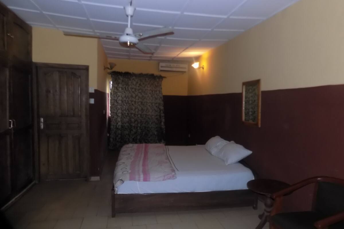 A bedroom with a bed, a ceiling fan, an air conditioning unit, a wooden door, and a chair.