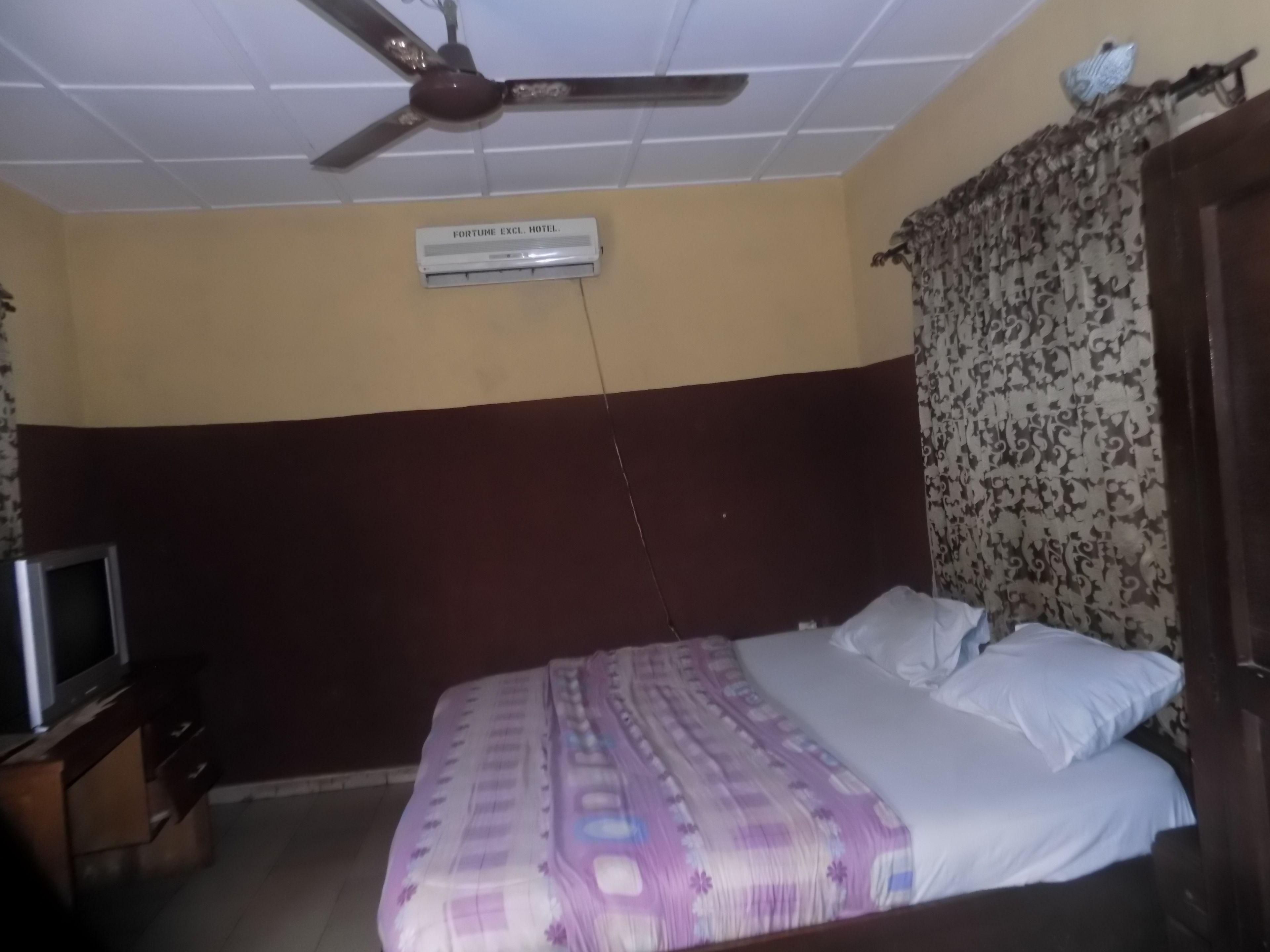 A hotel room with a bed, a ceiling fan, an air conditioning unit, a television, and a patterned curtain.