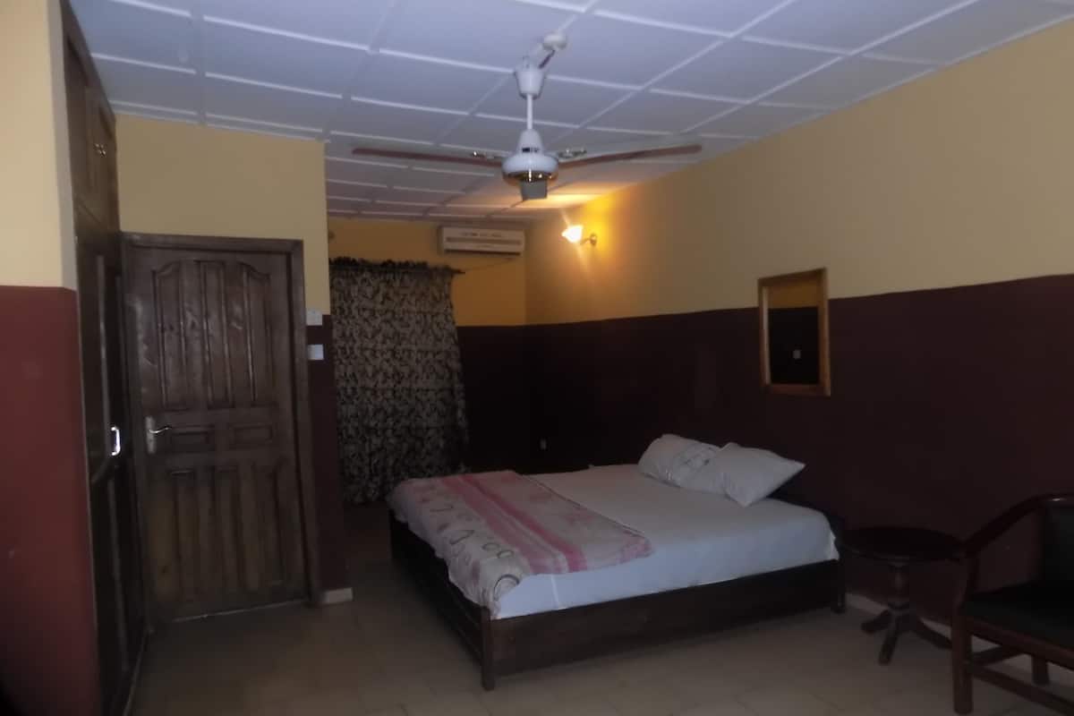A bedroom with a bed, a ceiling fan, an air conditioning unit, a wooden door, and a chair.