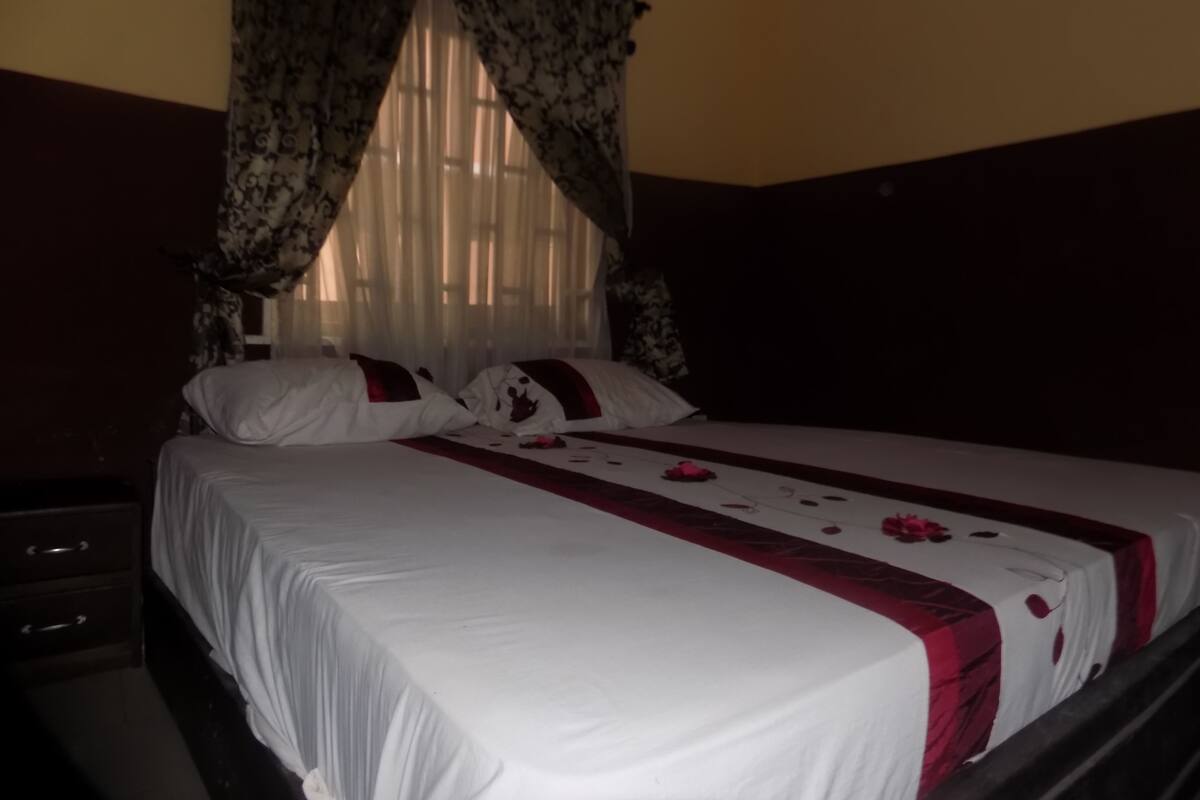 A bed with white bedding and a red and white floral pattern, a nightstand, and a window with sheer curtains.