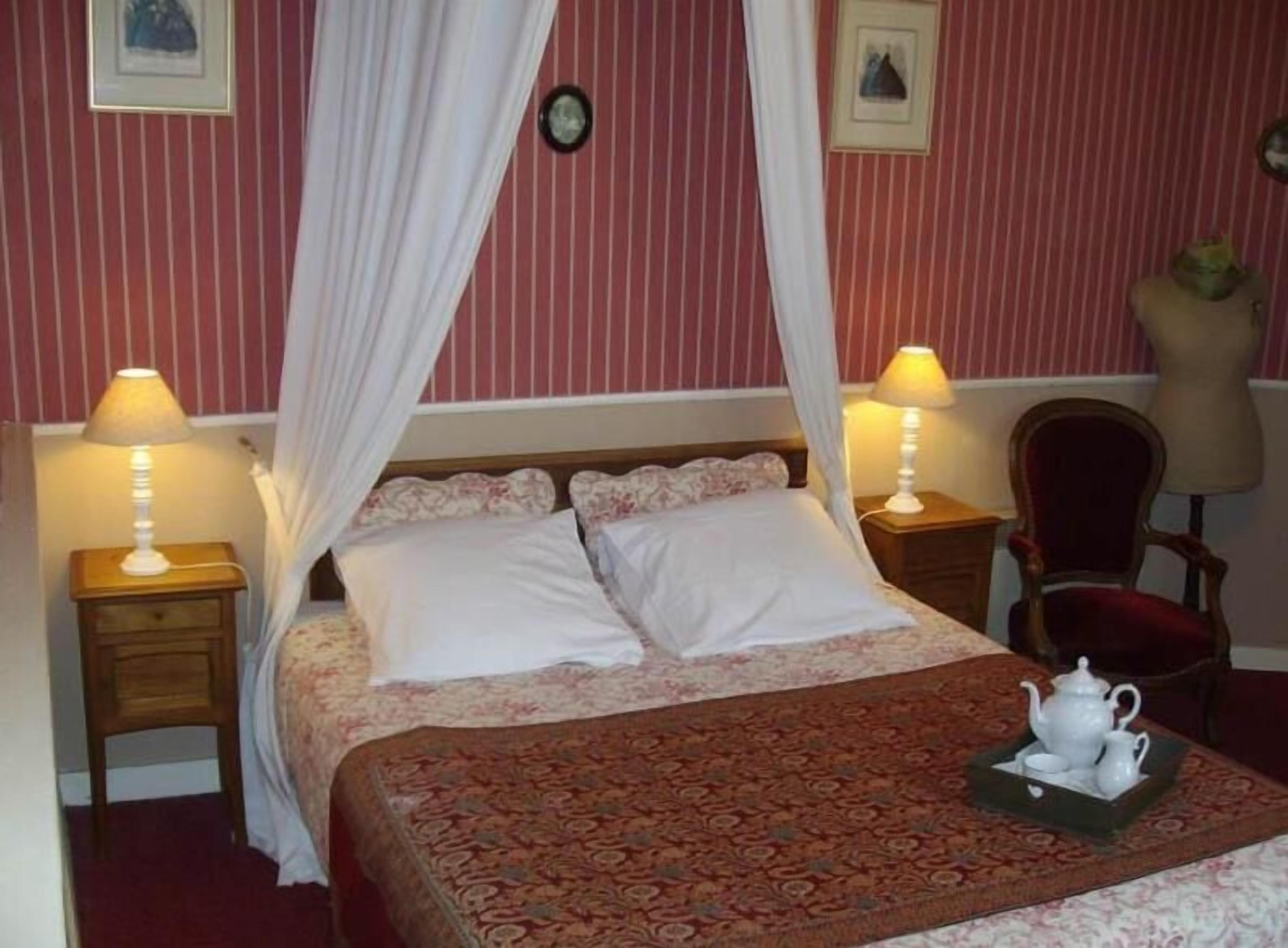 Un dormitorio con cama con dosel, dos mesitas de noche con lámparas, una silla y una bandeja con tetera y taza.