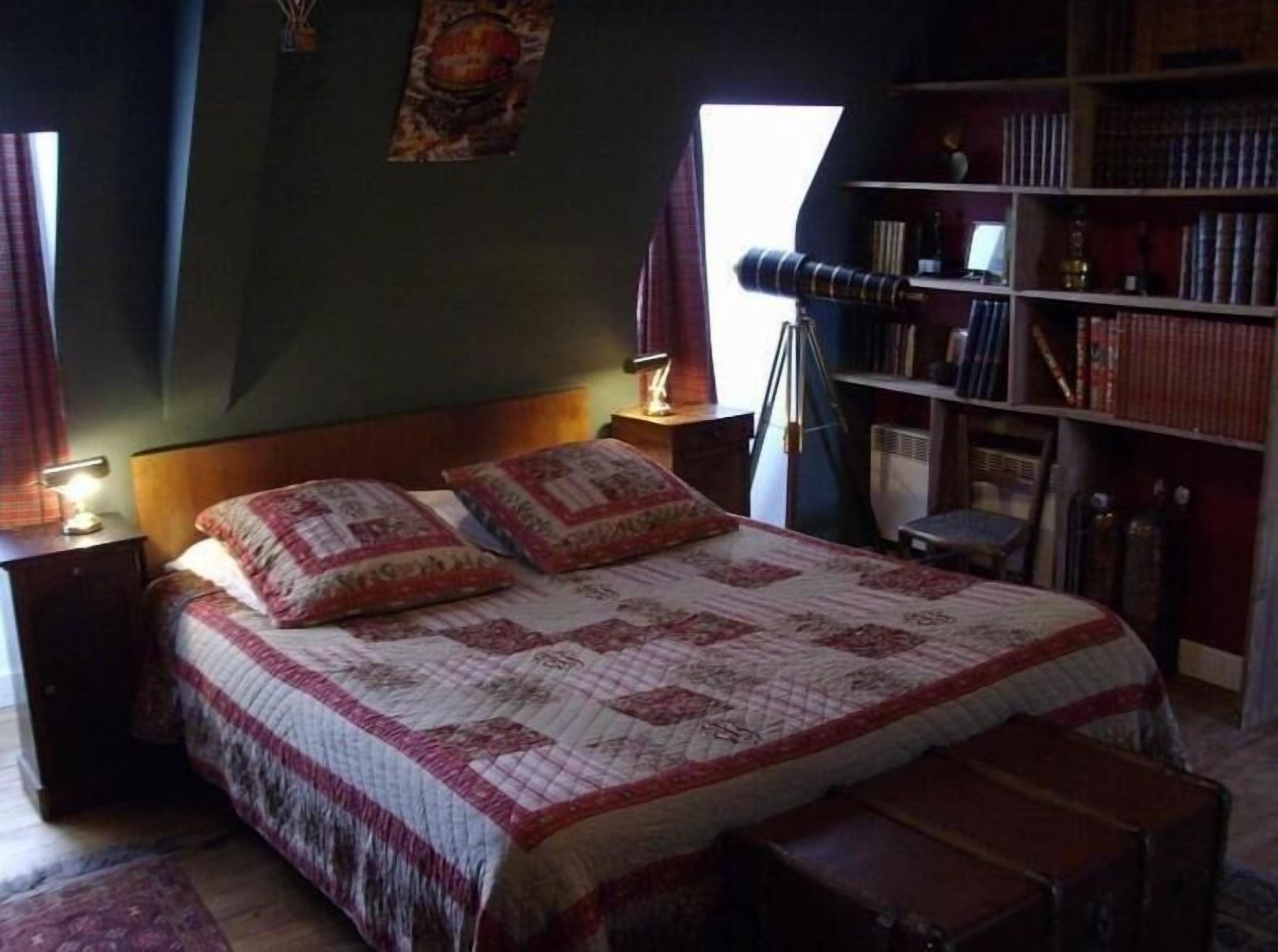 A bedroom with a large bed, a bookshelf, a telescope, and a window.