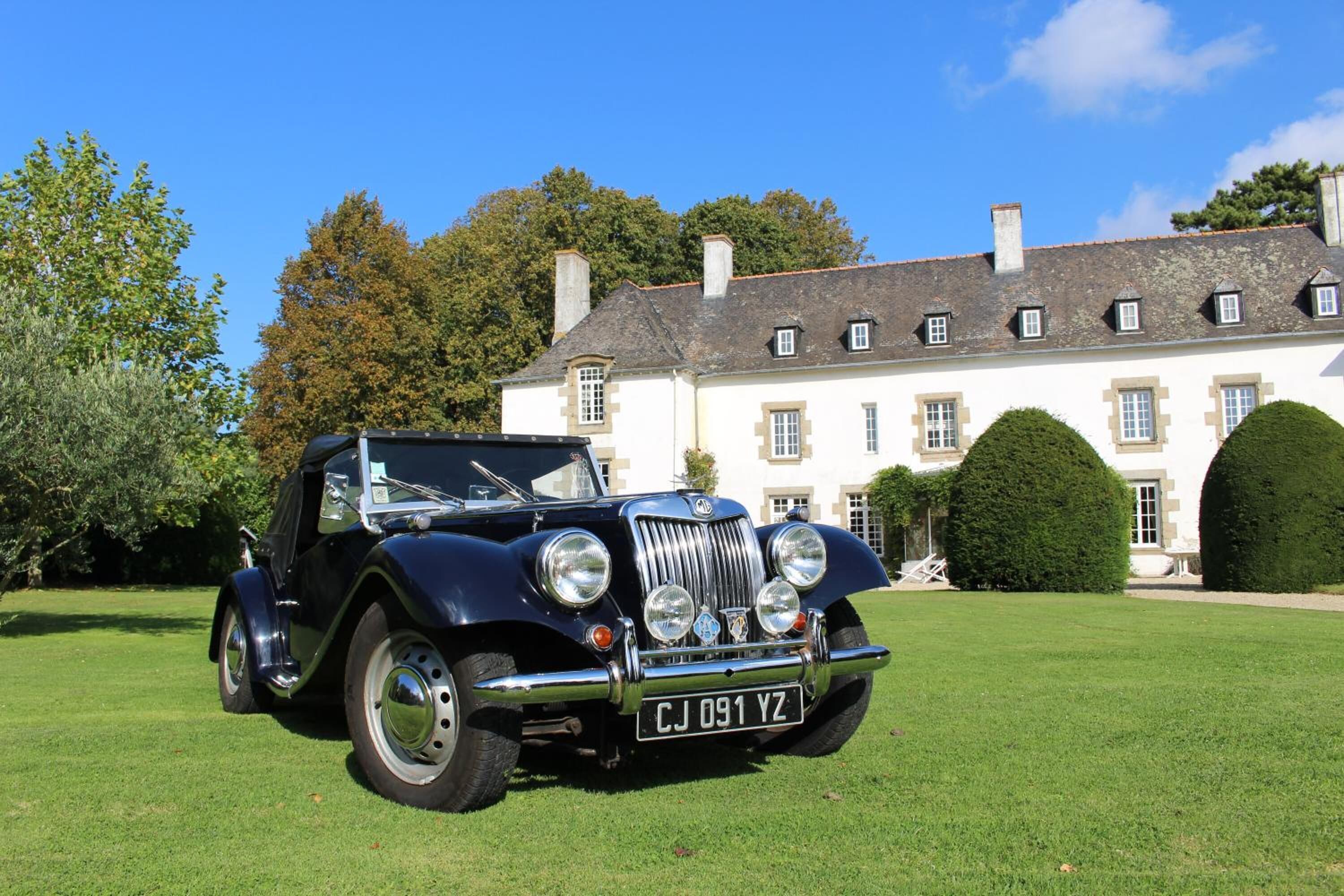 Une voiture ancienne est garée devant un grand bâtiment blanc, dont la façade est symétrique et qui possède de nombreuses fenêtres.