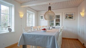 Een eetkamer met een lange tafel, witte stoelen, een fruitschaal en hanglampen.