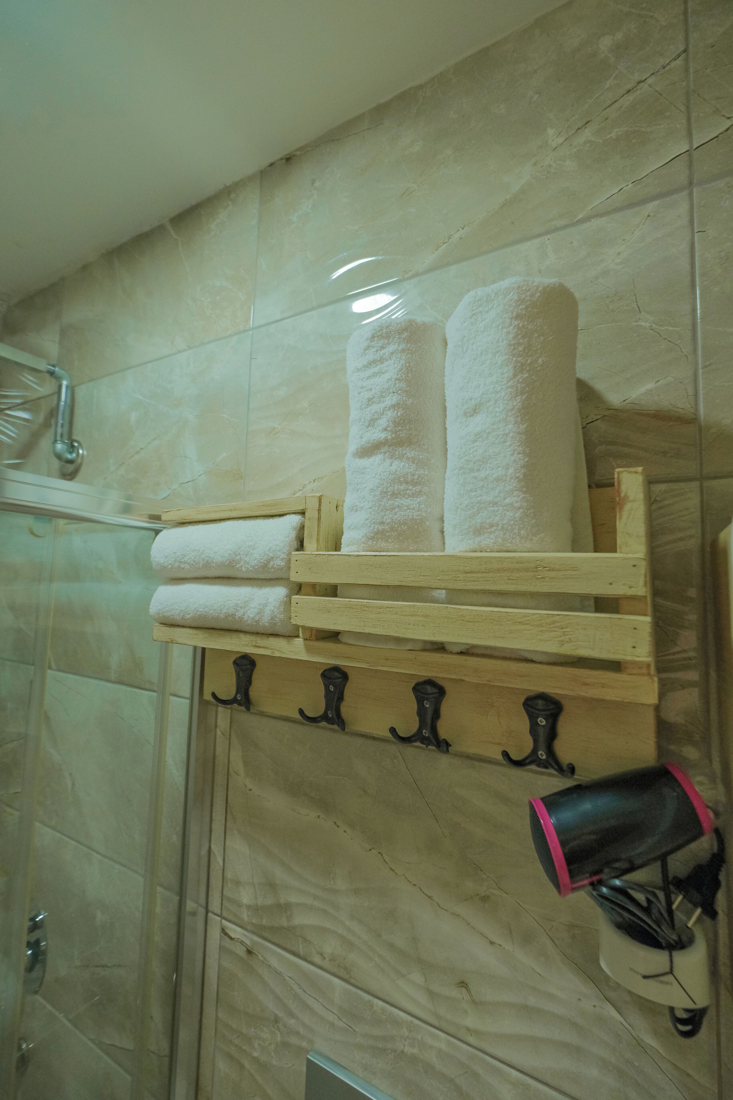 Un baño con pared de mármol, un perchero de madera con dos toallas enrolladas y un secador de pelo sobre un soporte blanco.