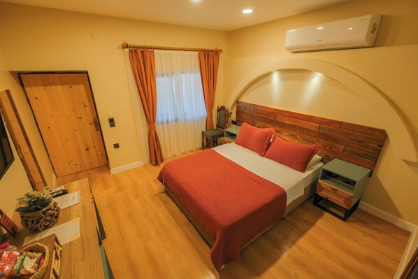 A bedroom with a wooden floor, a bed with red bedding, a nightstand with drawers, a chair, a window with curtains, and an air conditioning unit on the wall.