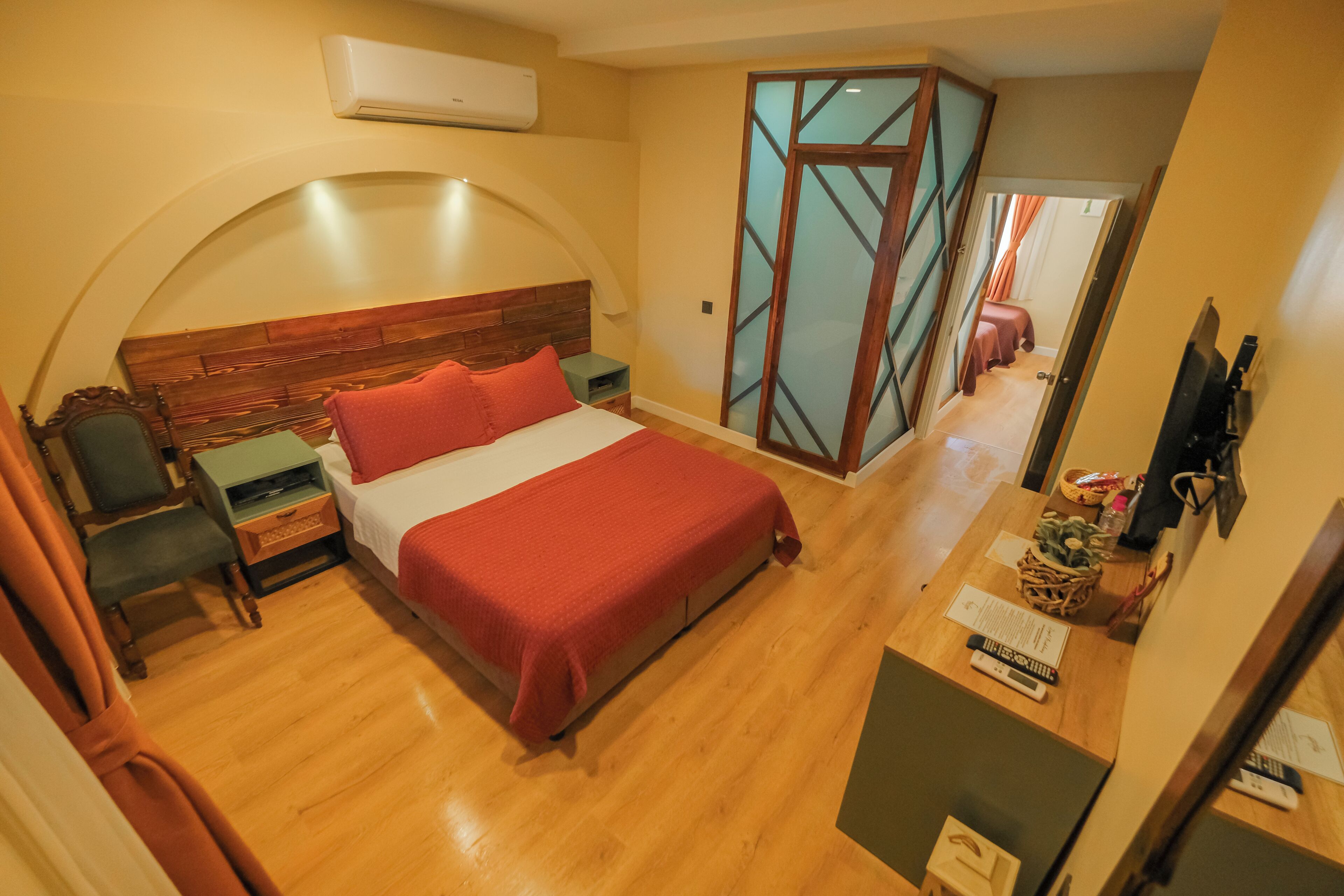 Un dormitorio con cabecera de madera, cama con ropa de cama roja, una mesita de noche, una silla, un escritorio con una cesta y un televisor.