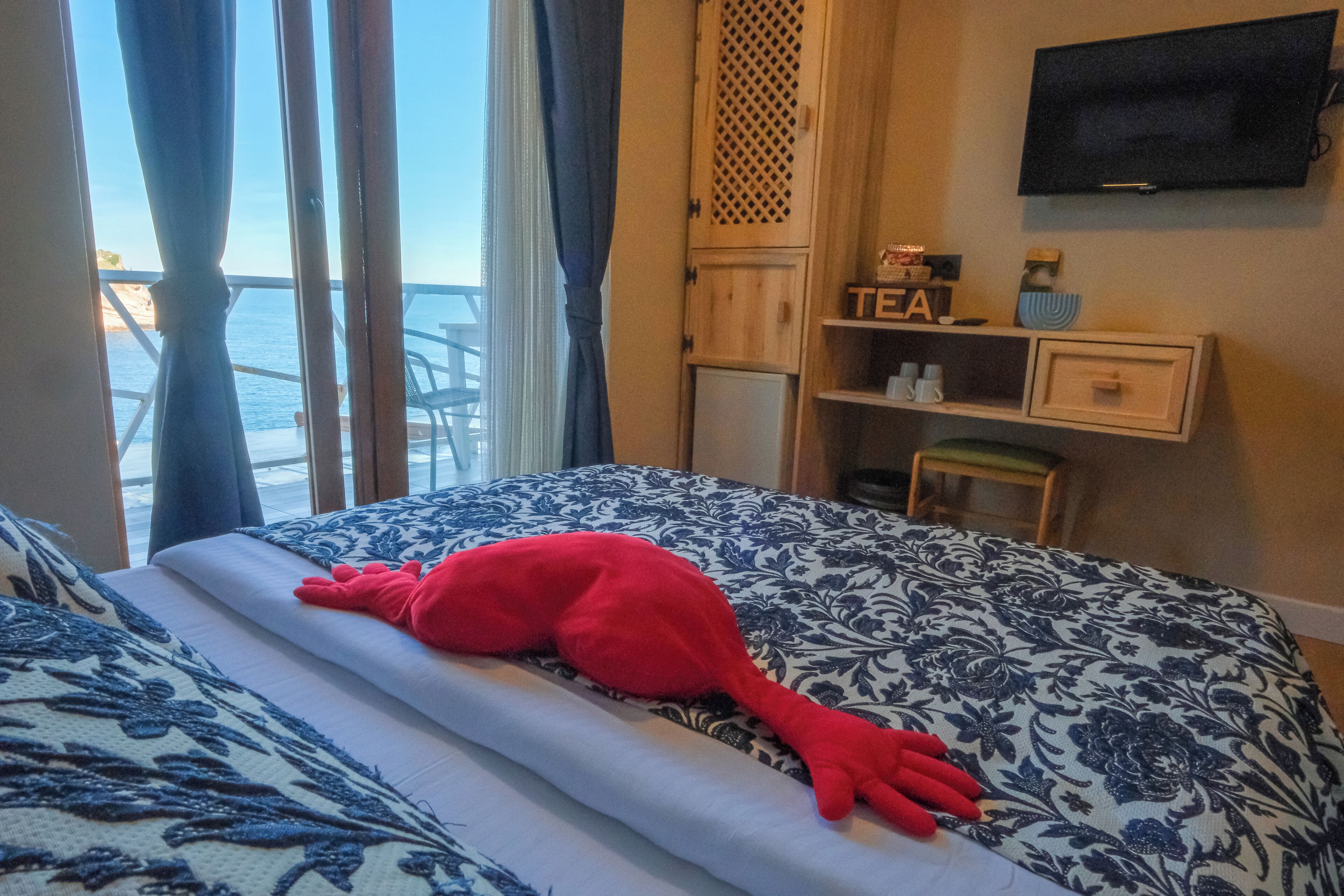Ein Hotelzimmer mit Bett, einem an der Wand befestigten Fernseher, einem kleinen Schrank mit der Aufschrift 'TEA' und Meerblick durch das Fenster.