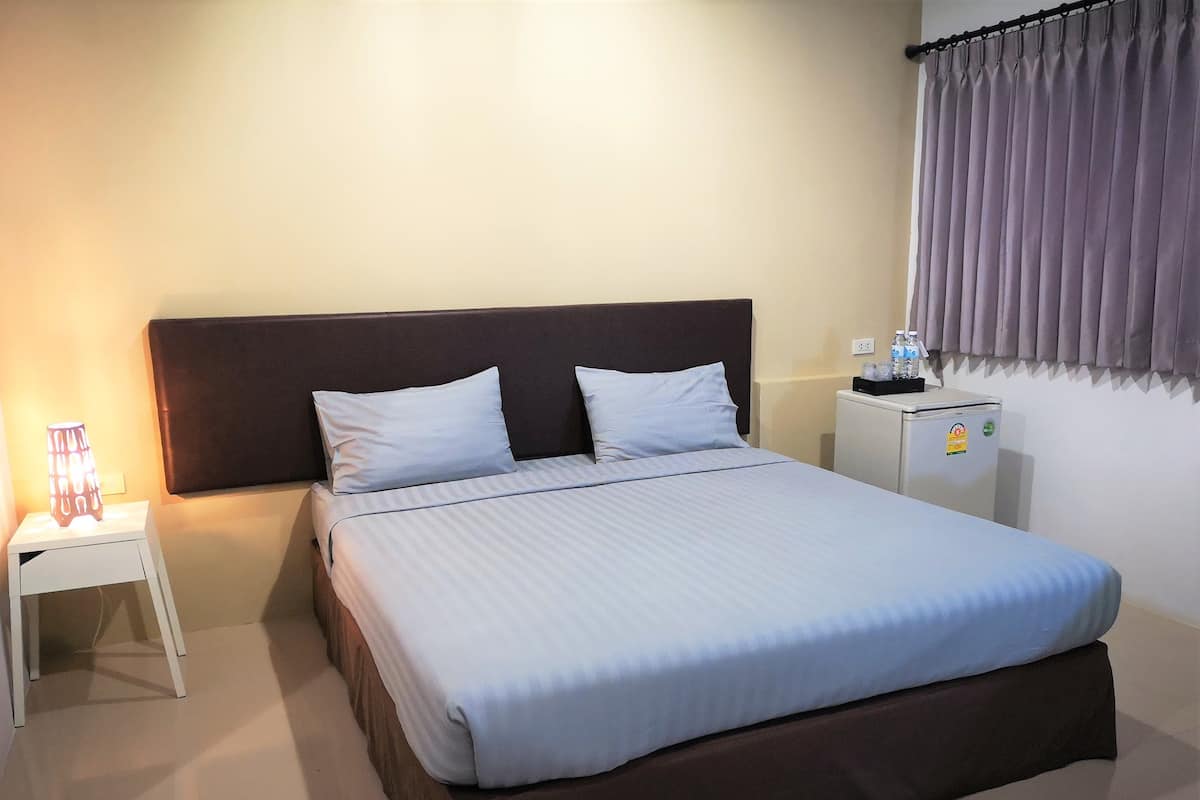 A neatly made bed with white bedding, a dark headboard, a bedside table with a lamp, and a small refrigerator on the right.