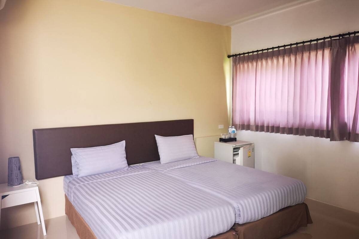 A double bed with a headboard, a bedside table, a nightstand, a window with curtains, and a small desk with a chair.