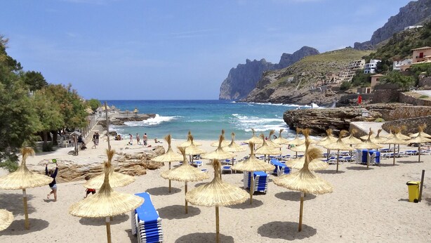 Ein Strand mit Reetsonnenstühlen und blauen Liegen, felsigen Klippen und Gebäuden im Hintergrund.