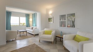 Ein modernes Wohnzimmer mit weißen Sofas, einem Couchtisch und einem Teppich. Es gibt Fenster mit Vorhängen, ein wandmontiertes Regal mit Dekoration und ein Gemälde.