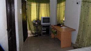 A small, compact room with a bed, a desk with a computer and a fan, and a television. - Royal Green Guest House (Abeokuta)
