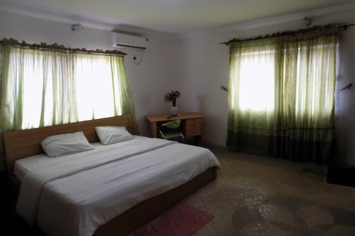 A bedroom with a bed, a desk, and a chair. There is a window with curtains and an air conditioning unit on the wall.