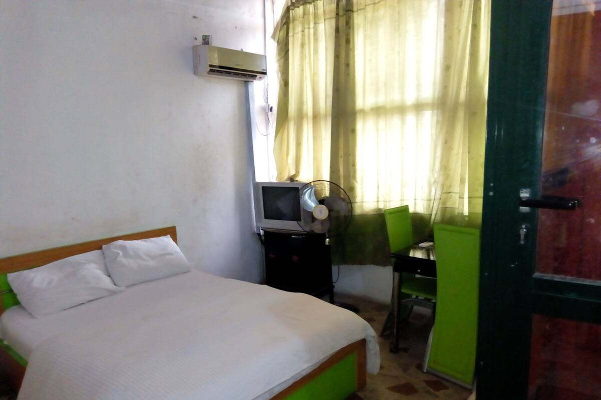 A room with a bed, a desk with a chair, an air conditioning unit, and a door.