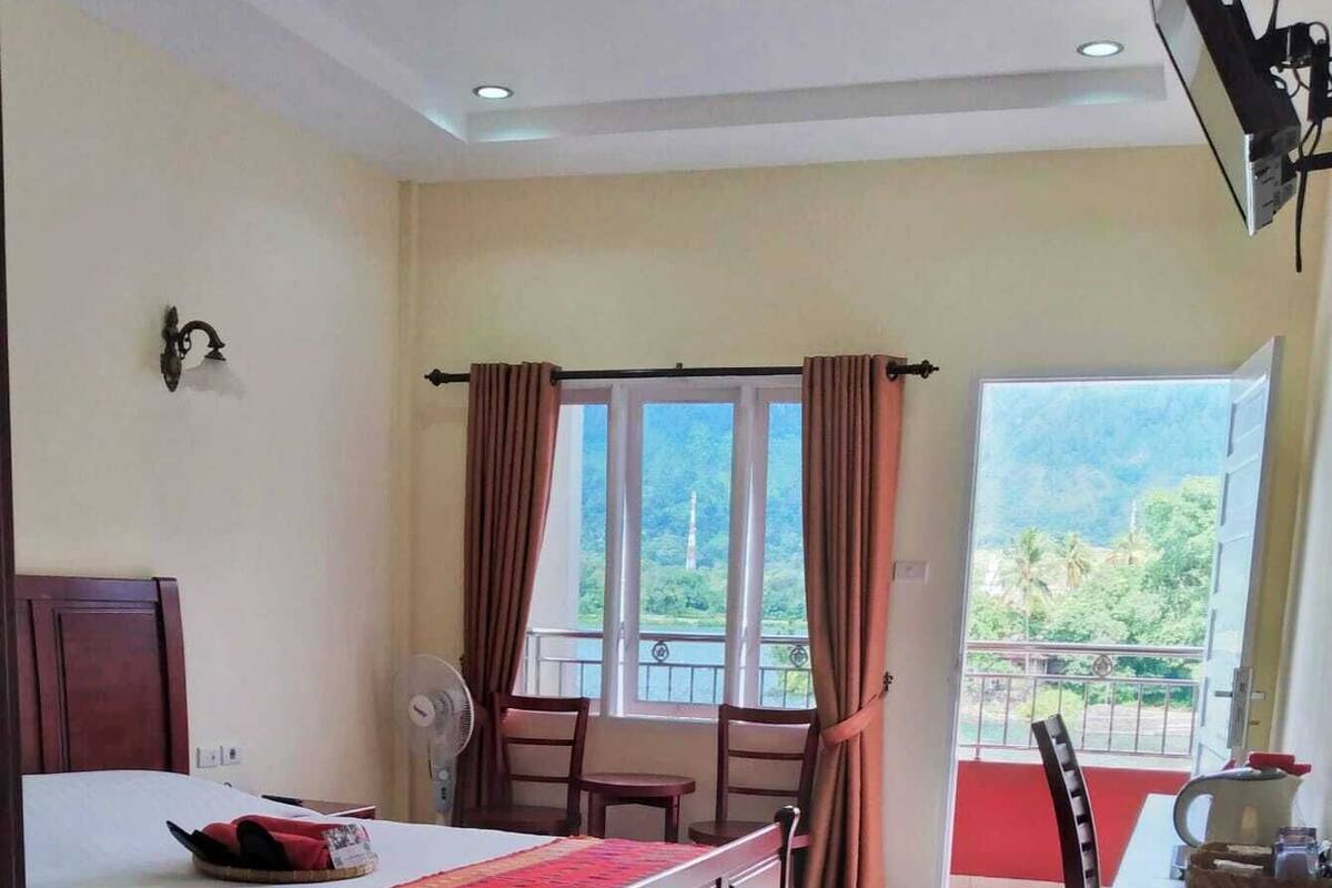 A hotel room with a bed, a dining table, a chair, a fan, and a window with curtains.