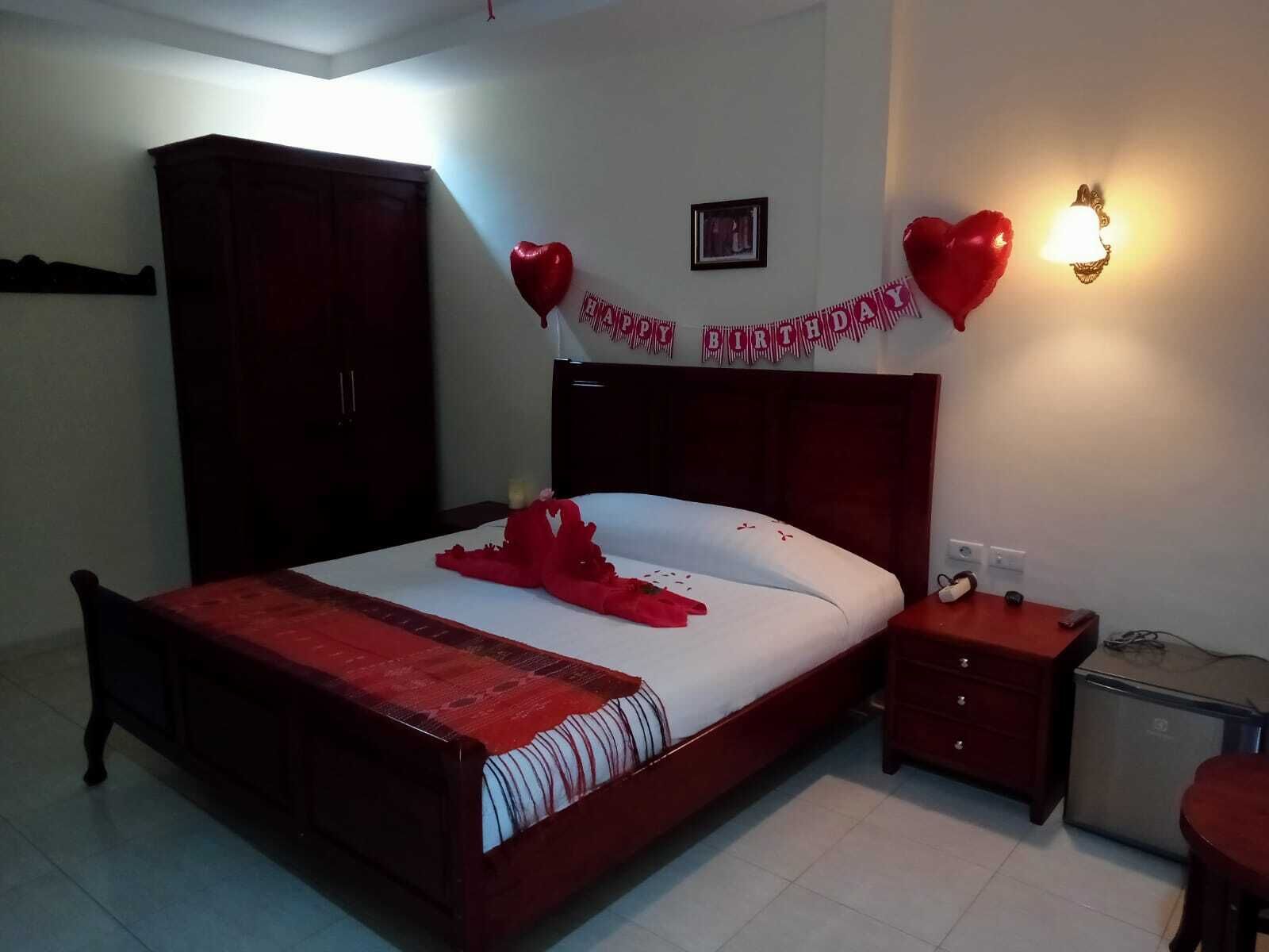 A bedroom with a wooden bed, a nightstand, a desk, and a chair. There is a 'Happy Birthday' banner on the wall.