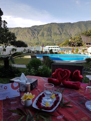 Una mesa dispuesta con un plato de postres, vasos y una decoración con forma de corazón, con vista a una piscina y un paisaje montañoso.