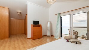 Quarto com cama grande, guarda-roupa de madeira, penteadeira, televisão e vista para o mar através de uma varanda.