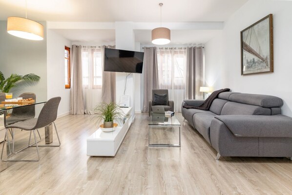 A modern living room with a grey sofa, a glass coffee table, a flat-screen TV, and a dining area with a glass table and chairs.