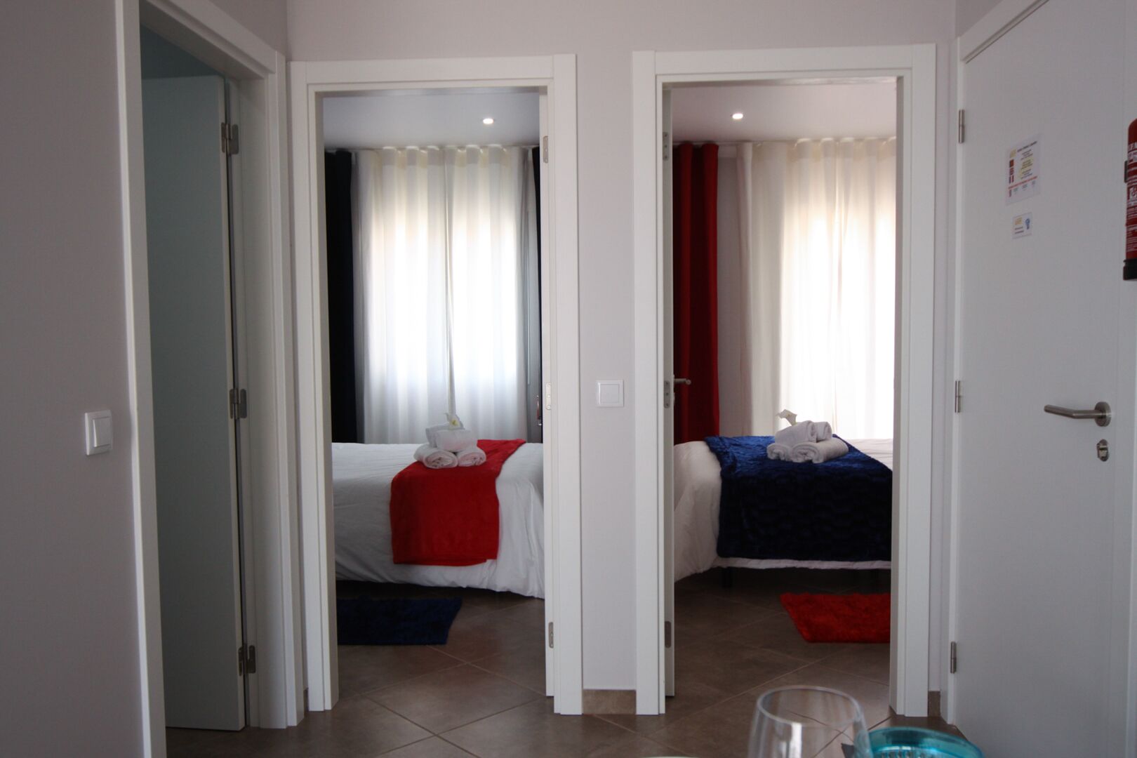 A hotel room with two beds, each with a red and blue blanket, and a white bedspread. There is a small blue trash bin and a white door with a handle.