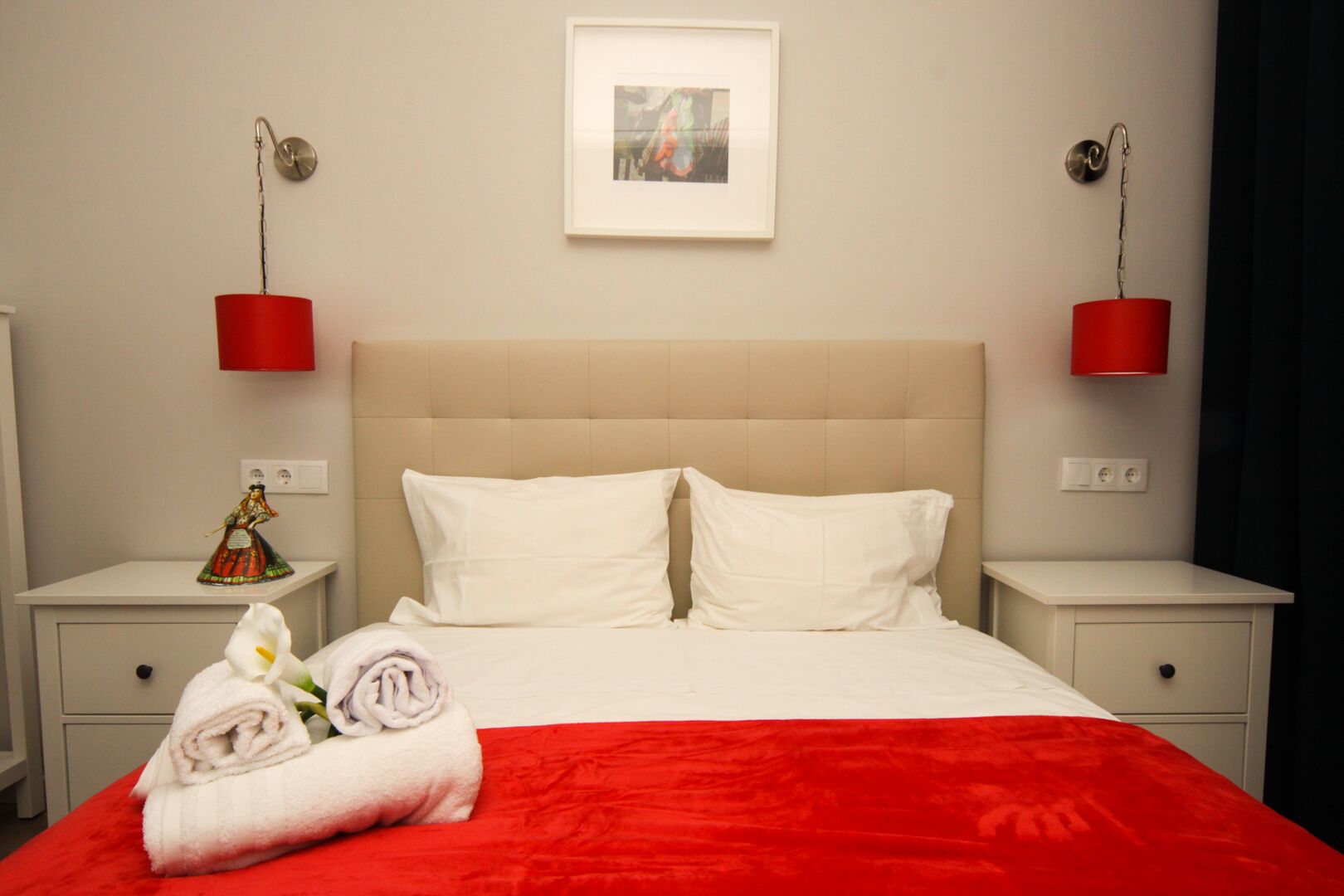 Ein Hotelzimmer mit einem Bett, zwei Nachttischen, einer roten Tagesdecke, einem weißen Kissen, einer roten Lampe, einem gerahmten Bild und einer kleinen Figur.