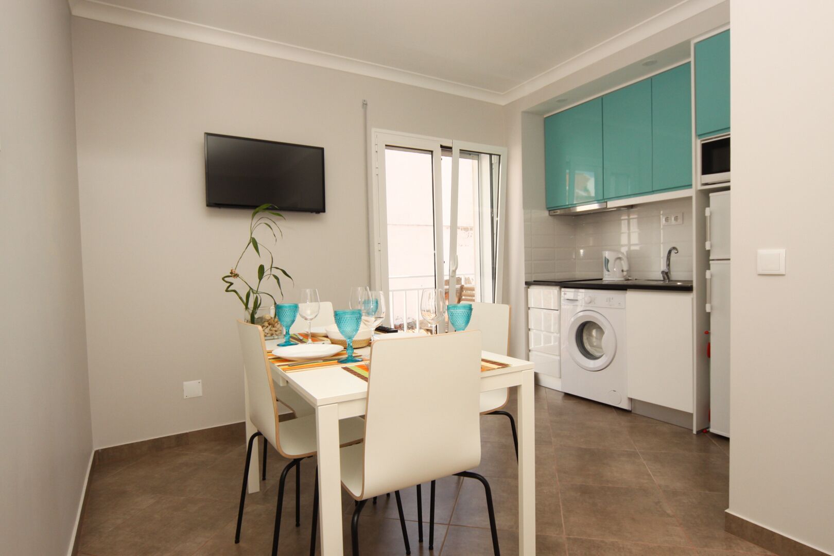 A compact kitchen with a washing machine, a dining area with a table set for two, and a TV mounted on the wall.