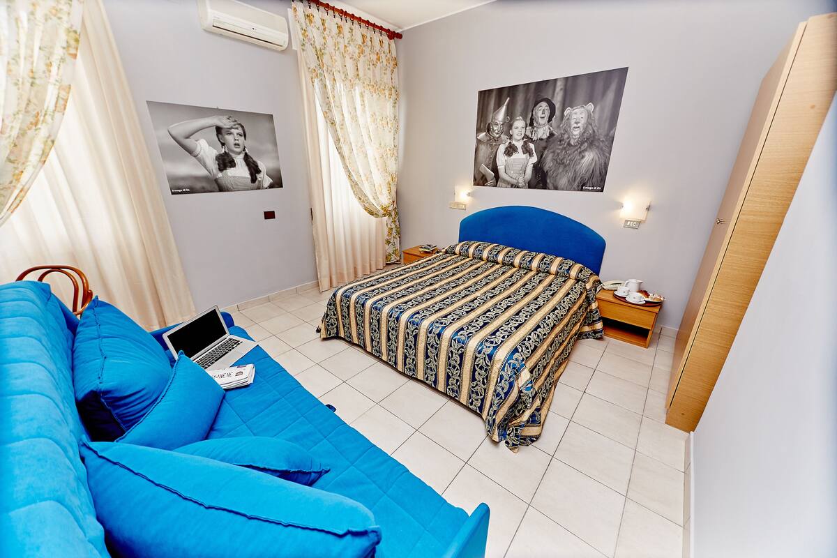 A bedroom with a blue sofa, a bed with patterned bedding, a laptop on the sofa, and two wall-mounted light fixtures.