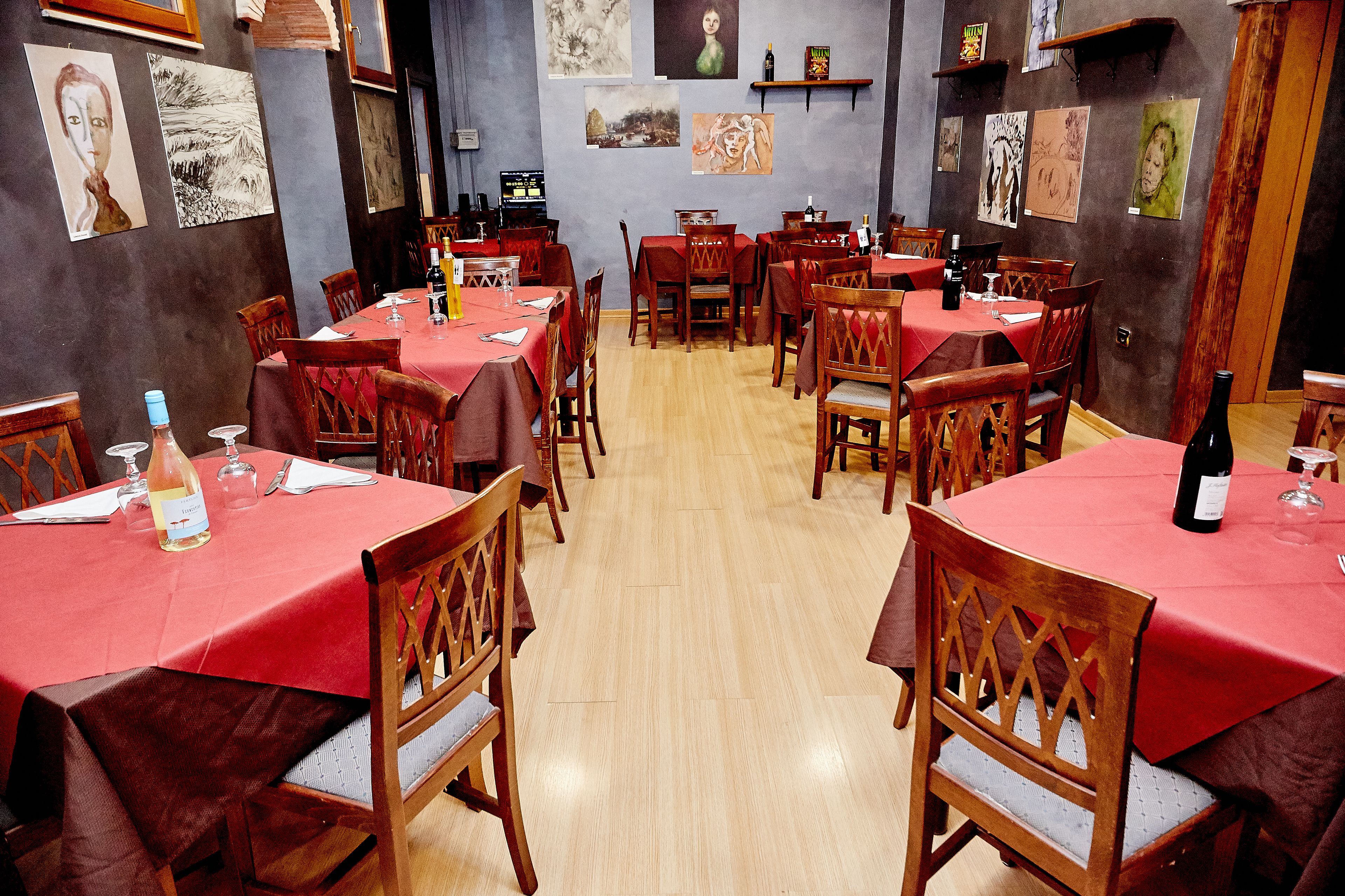 A restaurant with wooden tables and chairs, red tablecloths, and artwork on the walls.