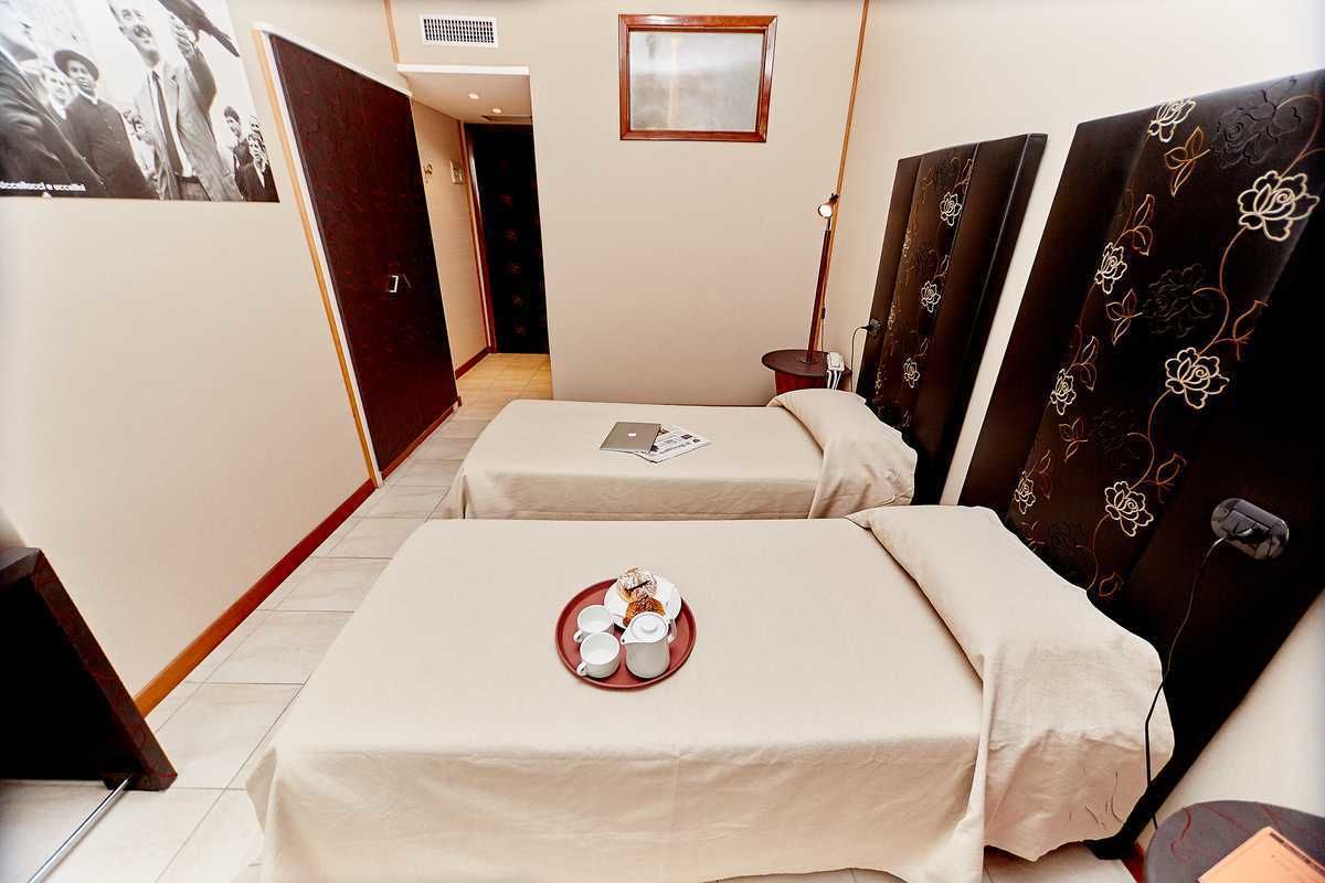 Een hotelkamer met twee bedden, een klein tafeltje met ontbijt, een foto aan de muur en een deur met bloemen gordijnen.