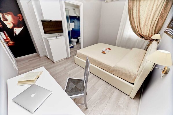 Une chambre d’hôtel avec un lit, un bureau avec un ordinateur portable, une chaise, une télévision et une salle de bain visible à travers une porte ouverte.