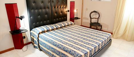 A bedroom with a tufted headboard, a patterned bedspread, a red nightstand, a desk lamp, and a chair.