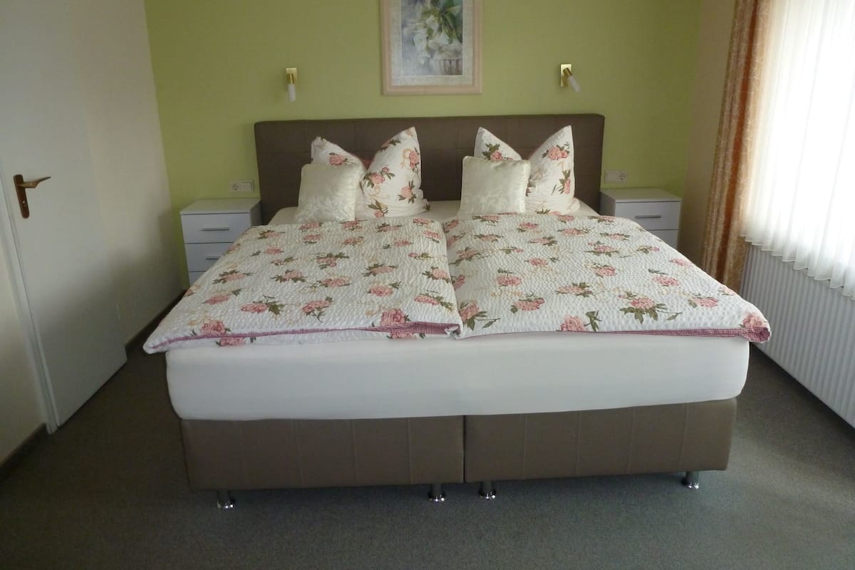 A double bed with floral bedding, two bedside tables, a nightstand, a door, and a framed picture on the wall.