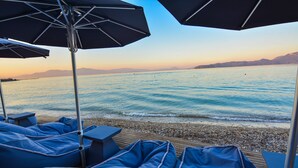 Un espace de détente aménagé sur la plage, avec des fauteuils bleus et des parasols, offrant une vue sur une mer calme et des montagnes lointaines.