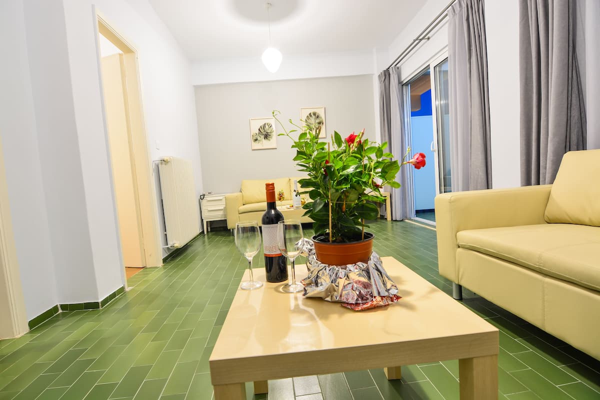 Een woonkamer met een groen betegelde vloer, een beige bank, een bijzettafel met een potplant en wijnkannen, en een radiator.