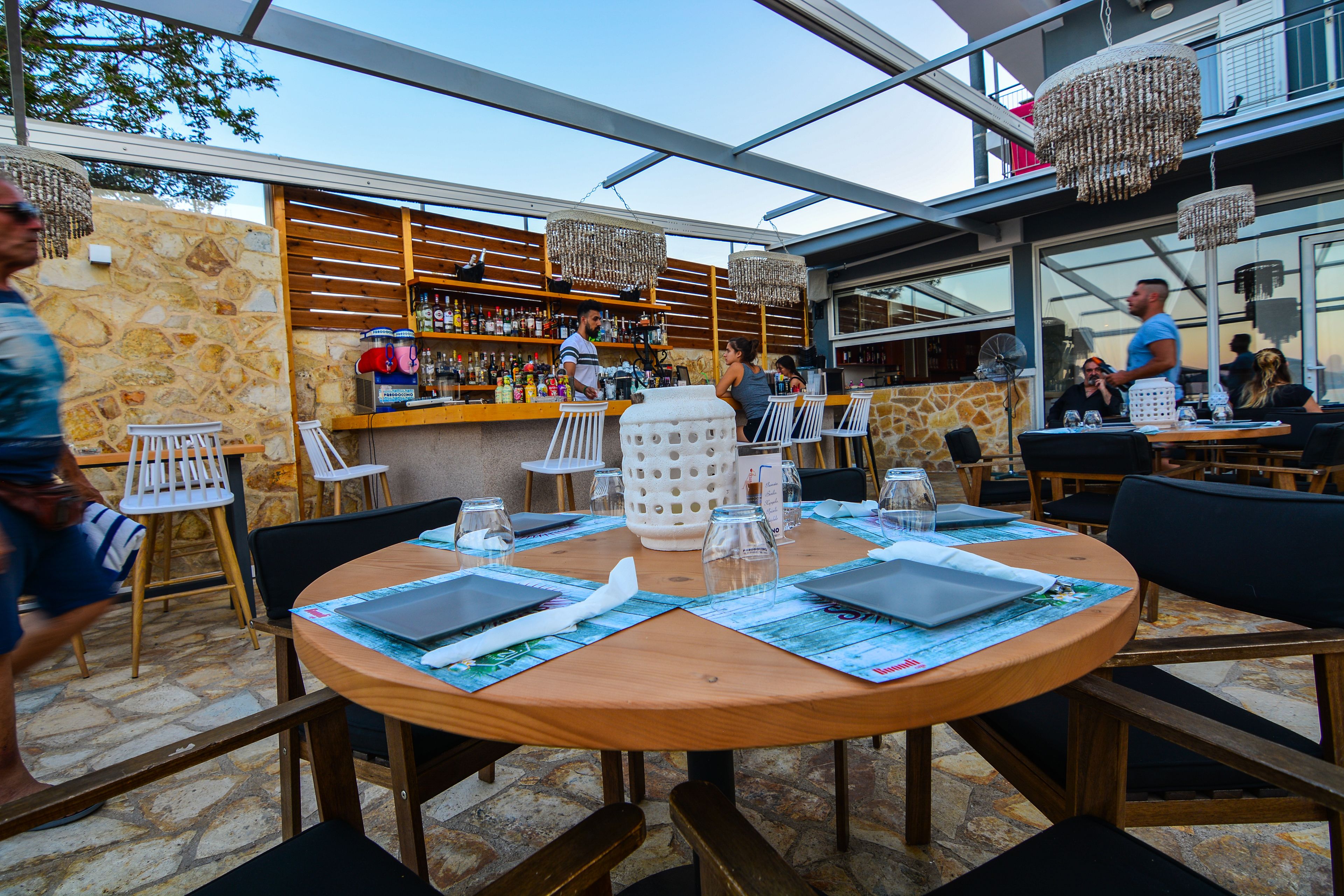 An outdoor dining area with round tables, chairs, and a bar with shelves of bottles.